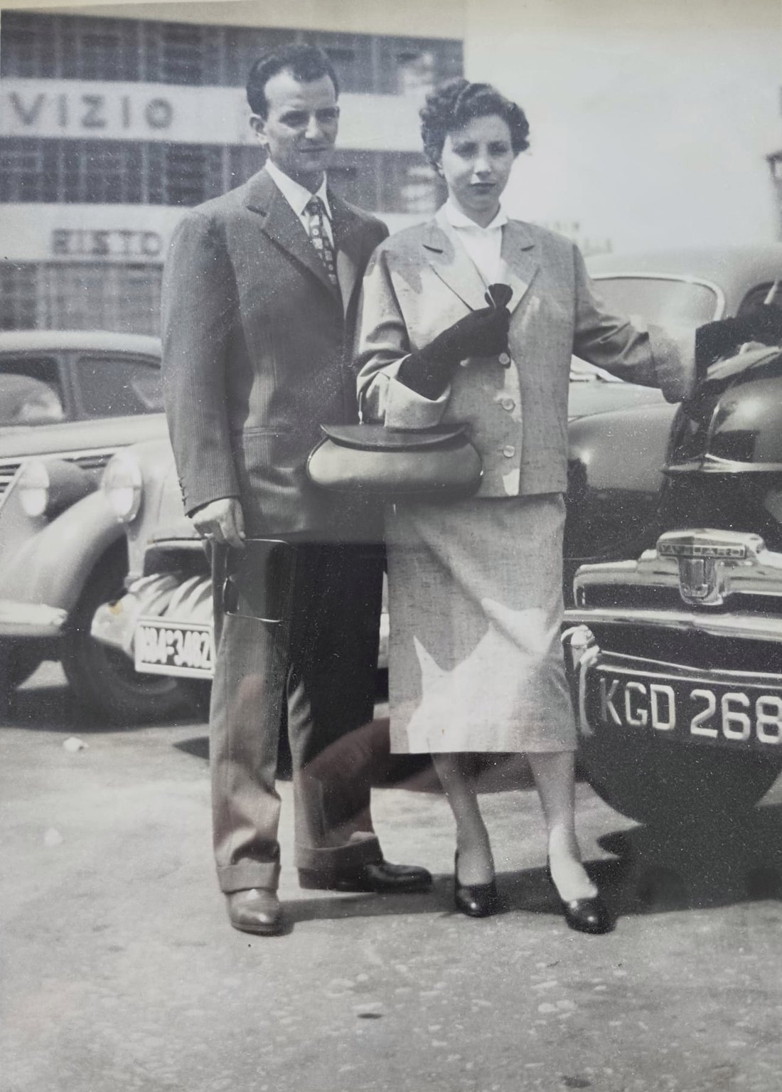 Leda e Bruno in abito elegante, davanti a due automobili degli anni Cinquanta.