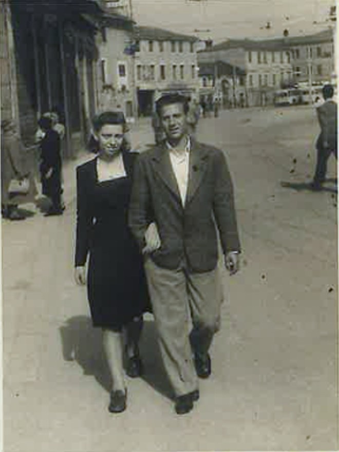 Wanda e Bruno a passeggio sotto braccio in una strada di citt&agrave;, con i portici a sinistra e una corriera sullo sfondo.